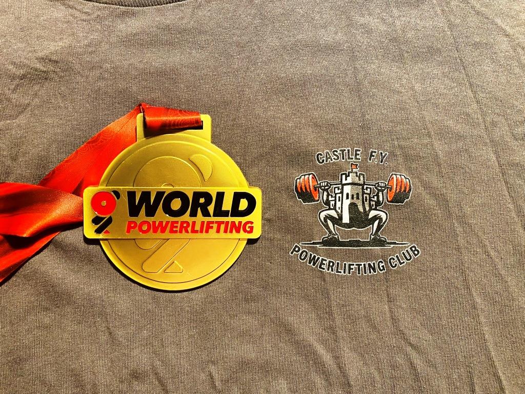 Close-up of a gold World Powerlifting Championship medal resting on a Castle F.Y. Powerlifting Club t-shirt. The contrast highlights the achievement and pride of the club, representing years of hard work and dedication.