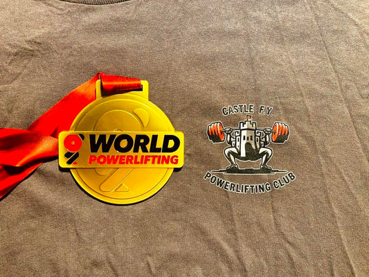 Close-up of a gold World Powerlifting Championship medal resting on a Castle F.Y. Powerlifting Club t-shirt. The contrast highlights the achievement and pride of the club, representing years of hard work and dedication.